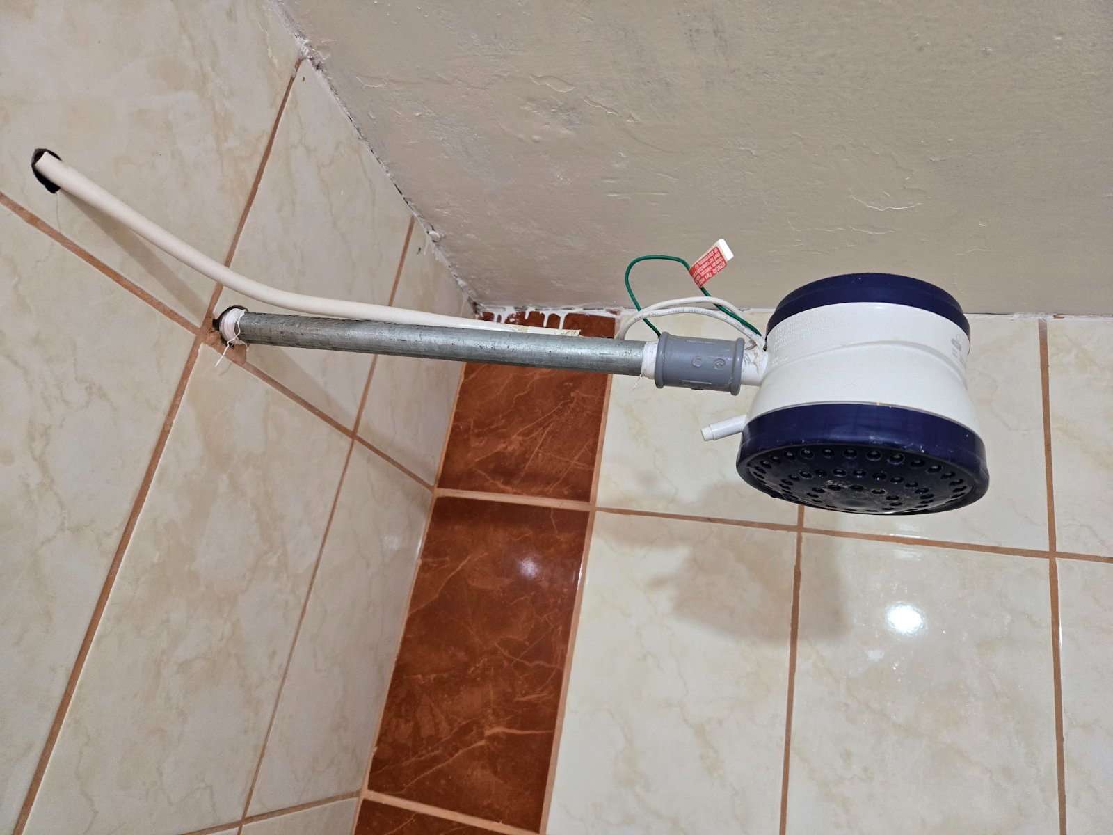 A 'suicide shower' or electric point-of-use heated shower head, with exposed wires, mounted on a tiled bathroom wall.